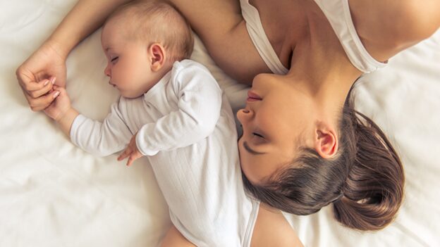 Tired Boynton Beach mom lying in bed with her baby, finally resting after struggling with sleepless nights and stress.