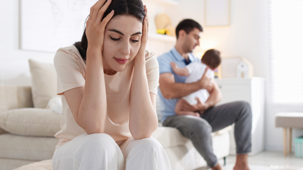 Overwhelmed new mother sitting with her head in her hands while her partner holds their baby in the background.