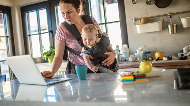 Burned-out working mom juggling remote work while holding her baby in a busy kitchen, looking exhausted and stressed.